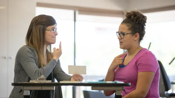 Two people are sitting at a desk. One is holding a card and pointing to their mouth. The other person is pointing to their throat.