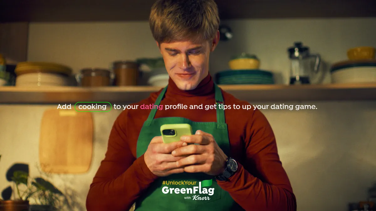 A man holds a phone while wearing a green apron and red turtleneck sweater.