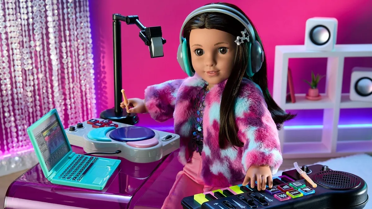 American Girl doll sits at a toy desk in front of a computer and a microphone.