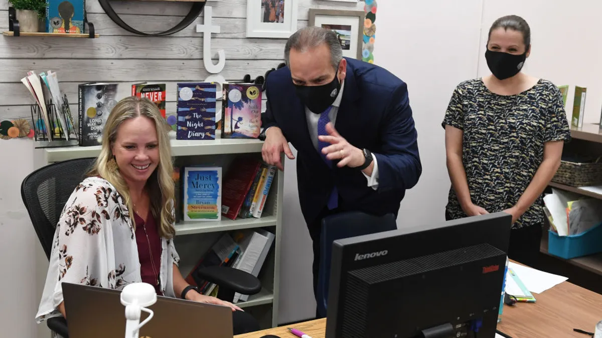 Three people are in a classroom. One person is sitting at a desk with an open laptop. The person in the middle is wearing a face mask and waving to the laptop. The third person is standing looking at the laptop.