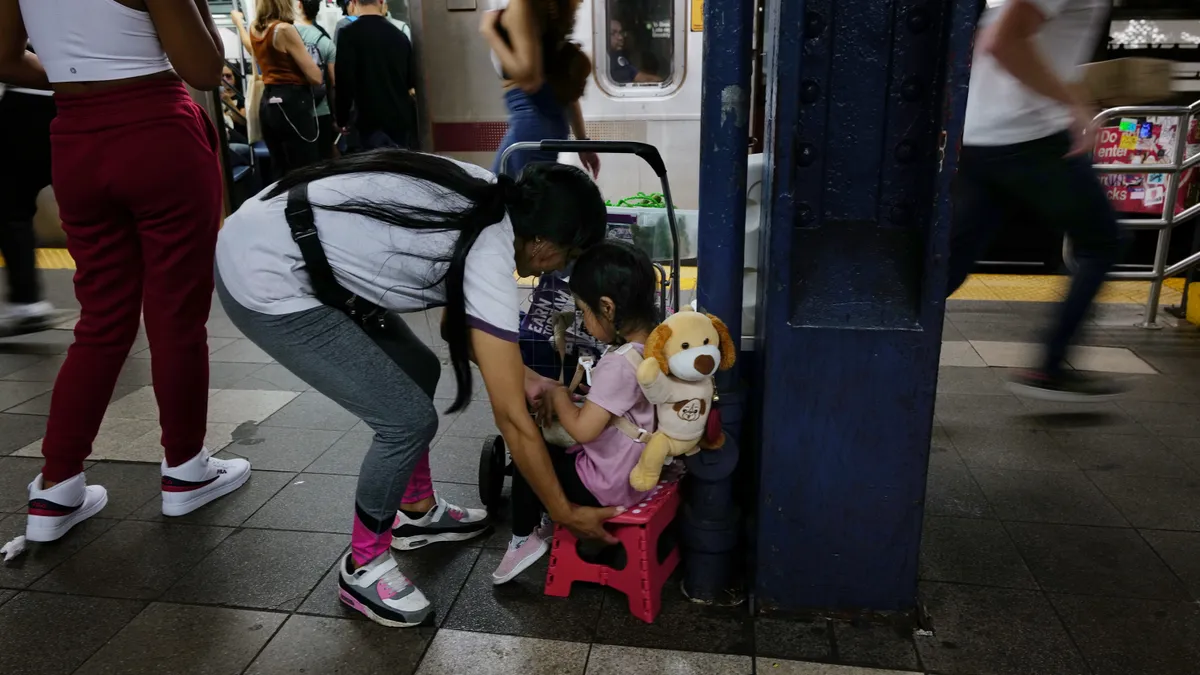 An adult leans down toward a seated child. Behind the child is a stuffed animal. They are in a subway station with other people walking past.