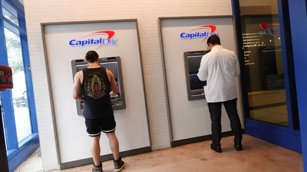 Two people use Capital One ATMs. A sign above them reads "Check your (inner) balance"