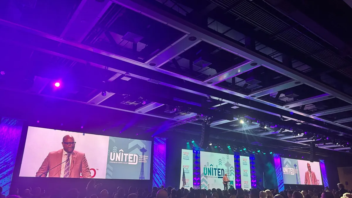 Three large screens appear above a stage that say "UNITED" with a person speaking who is displayed on the outer screens.