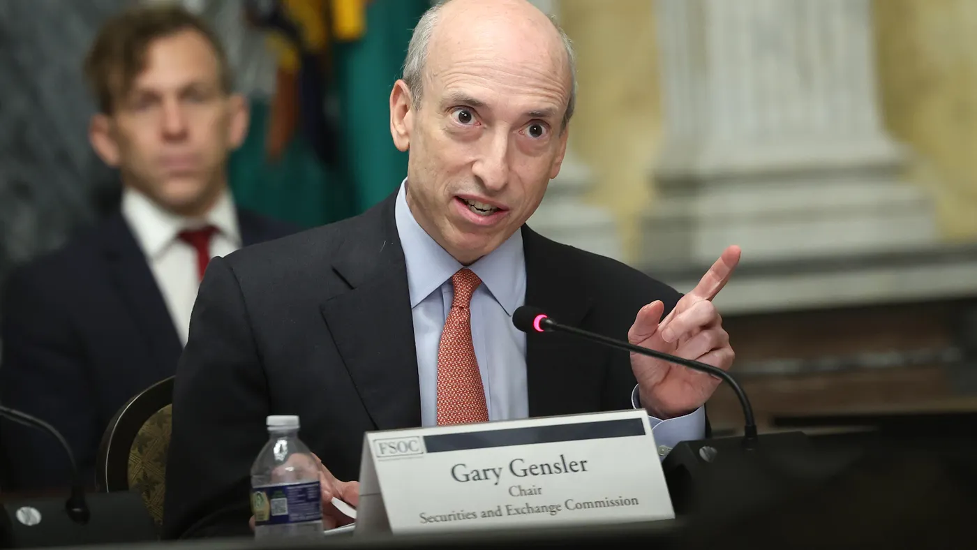 Gary Gensler is in focus speaking with a finger raised in front of a name card. An aide sits behind him out of focus.