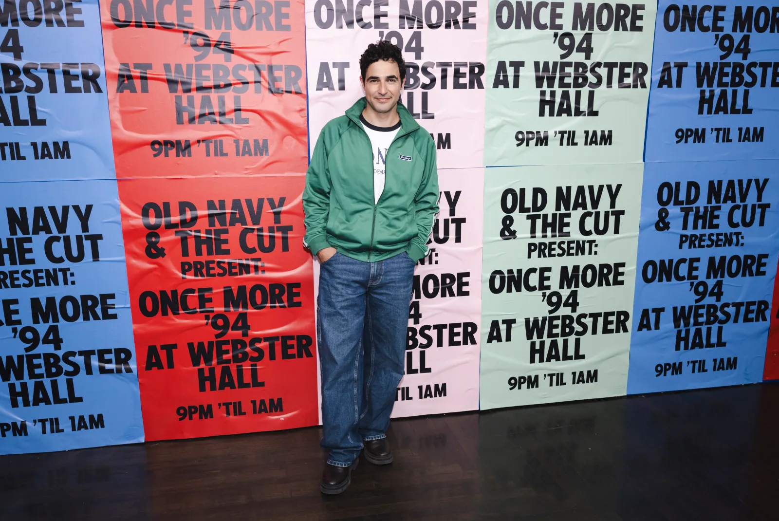 A [person in a track jacket and jeans stands against a colorful wall