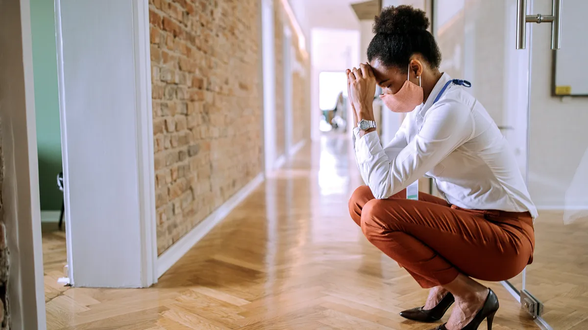 A masked Black person squats in distress in their workplace hallway