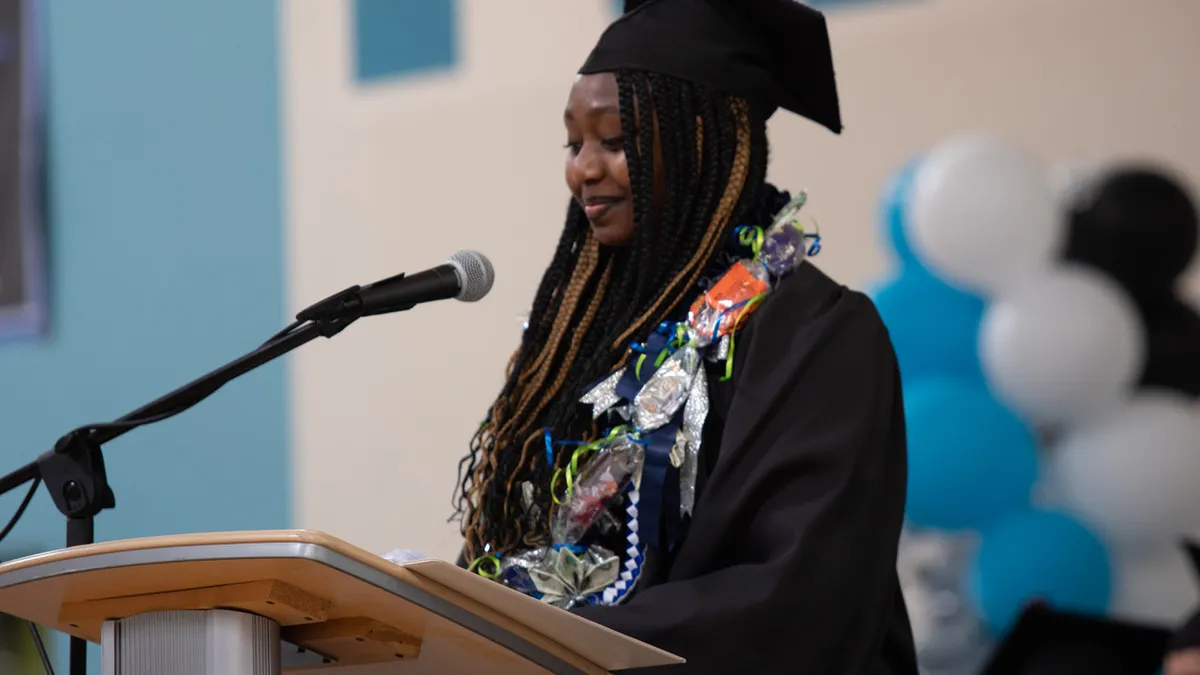 A Black student stands at a podium to deliver her remarks during a high school graduation ceremony. There are flowers around her neck and out-of-focus blue, white, and black decorations behind her.