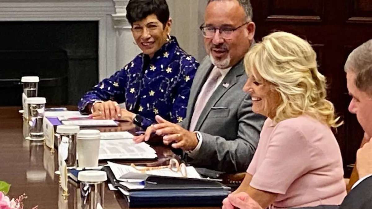 Four adults sit on one side of a conference table. Water glasses and notebooks are on the table and a fireplace is at the far end of the room.