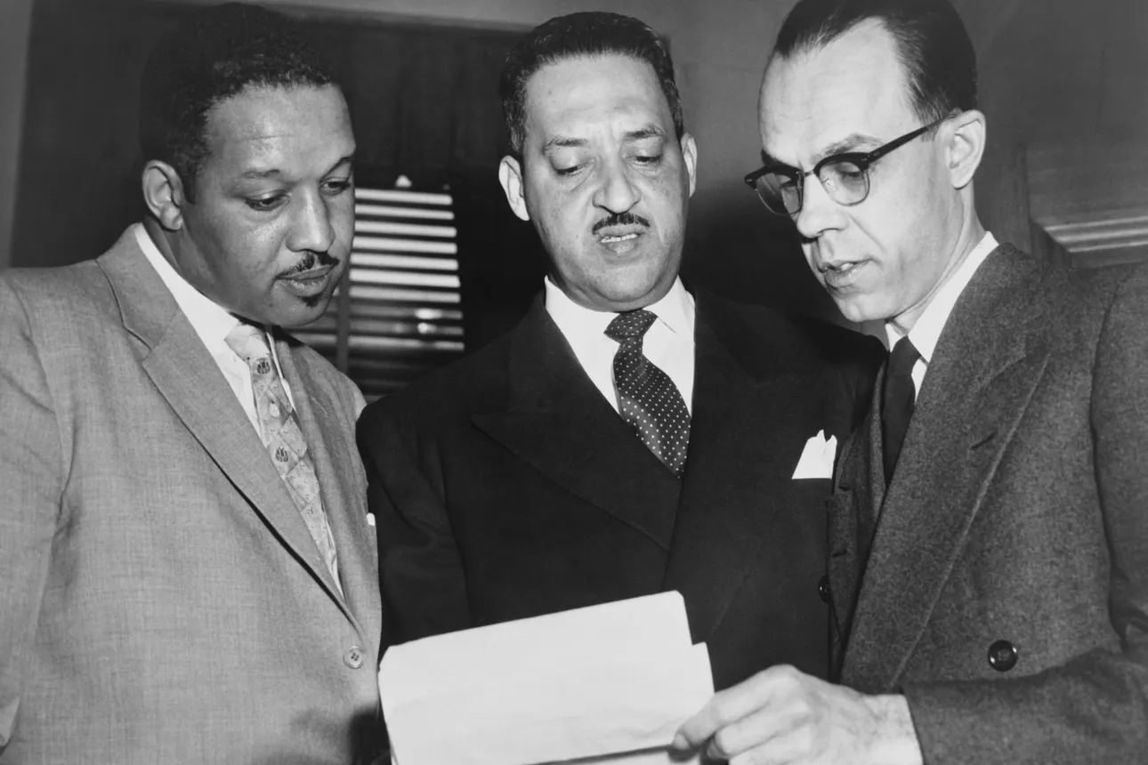 Three men dressed in suits examine a stack of papers