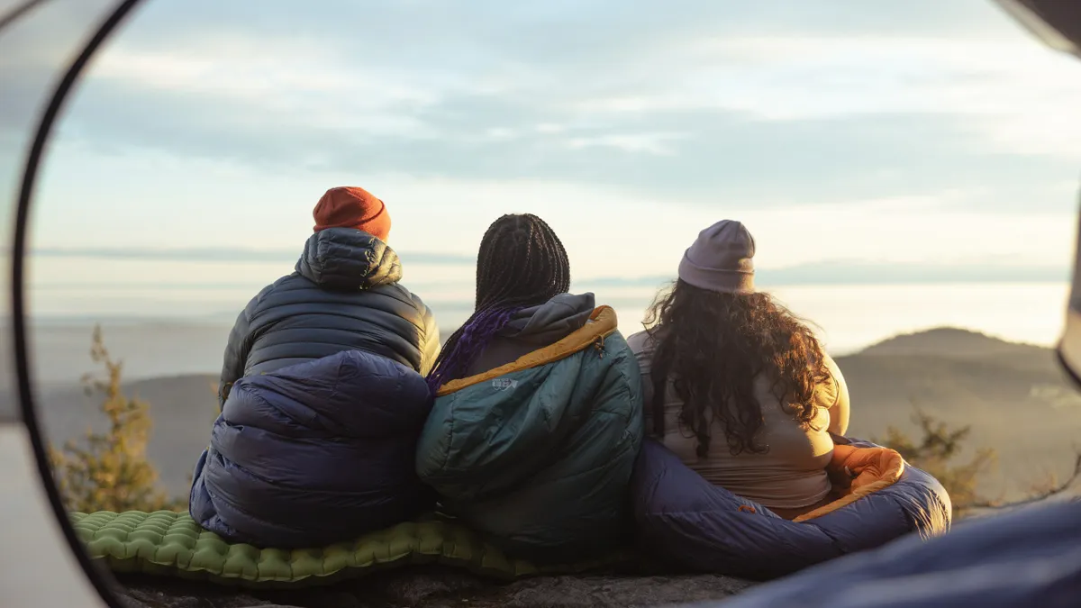 Three people sit outside in sleeping bags.