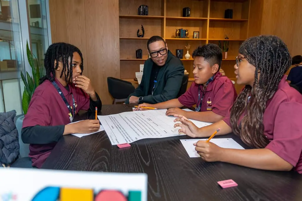 Three students and an adult are sitting at a table in a room. The students have pieces of paper on a table and are holding pencils