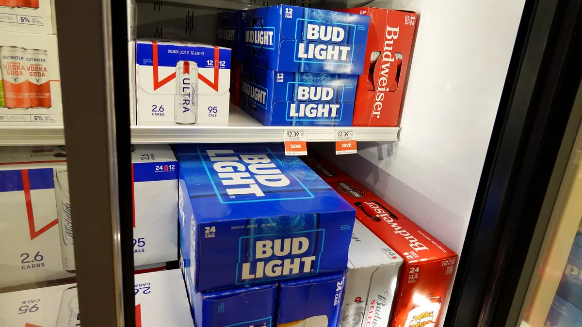 Cardboard cases of Bud Light cans sit on shelves in a grocery store refrigerator case, alongside cases of Budweiser and Michelob Ultra