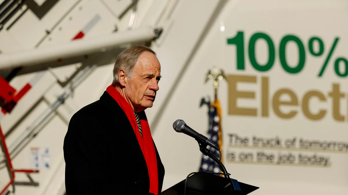 A man wearing a winter coat and scarf stands in front of a garbage truck with the words "100% Electric" printed on the side.