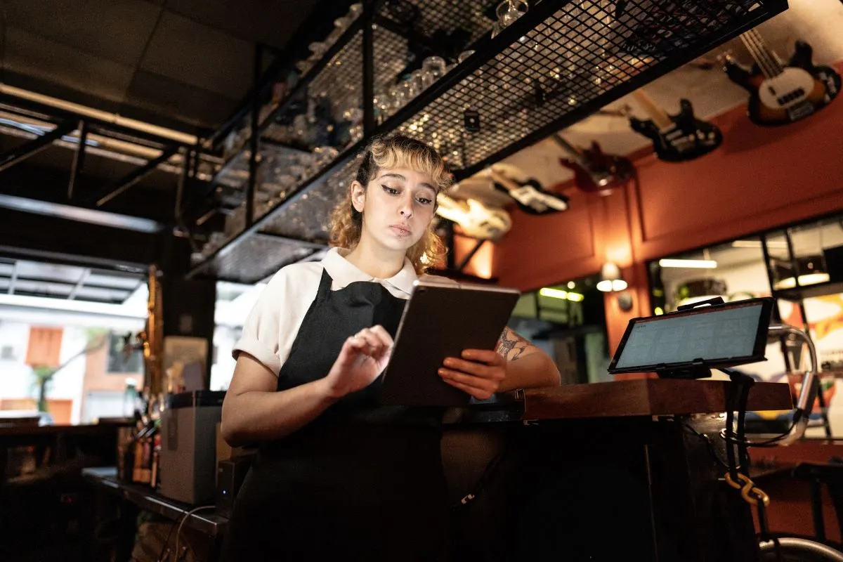 A female waitress wearing a black apron looks at a black tablet.