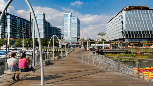 Two people sit on a swing on the left of the photo with a wooden boardwalk down the middle and large blue and white buildings in the background on a sunny day.