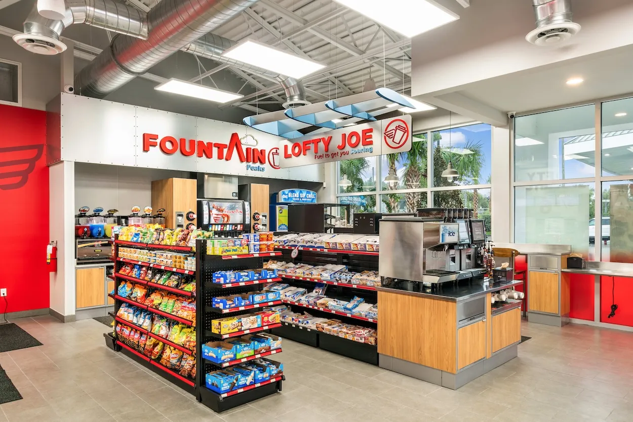 A photo of the interior of a store, showing the beverage dispensers as well as some of the front windows. A sign over several coffee dispensers says &quot;Lofty Joe&quot; while a sign over the soda and other cold drink dispensers reads &quot;Fountain peaks.&quot;