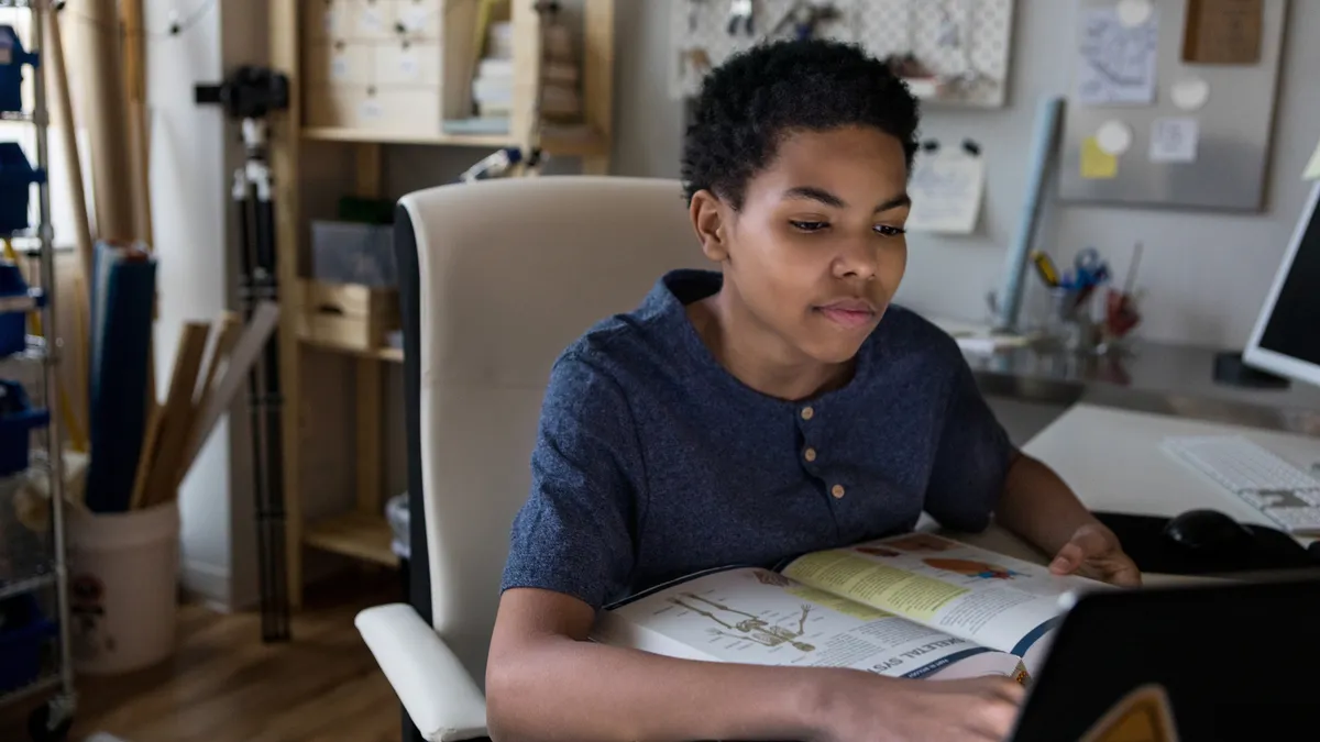 African American teenage boy uses laptop while studying for a biology test. A biology textbook is in front of him.