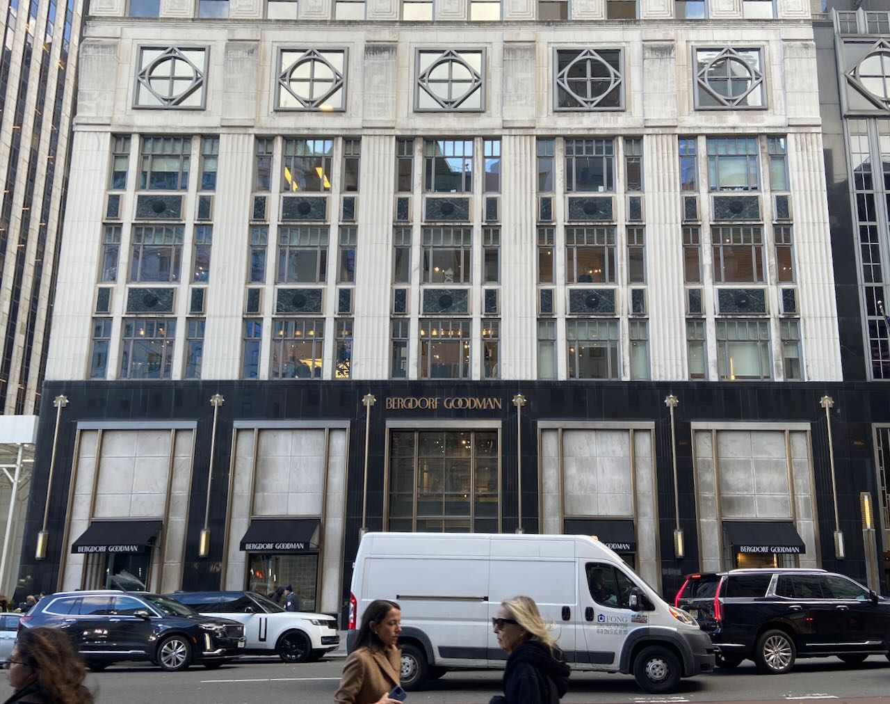 People and cars going past a big Art-Deco building.