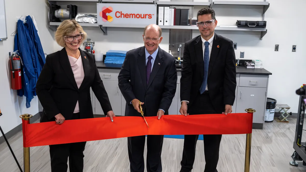 Three people wearing goggles and business suits holding a red ribbon.