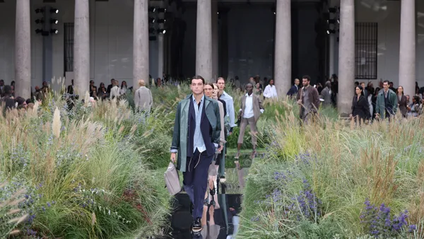 Models in suits walk between patches of tall grass on a fashion runway.