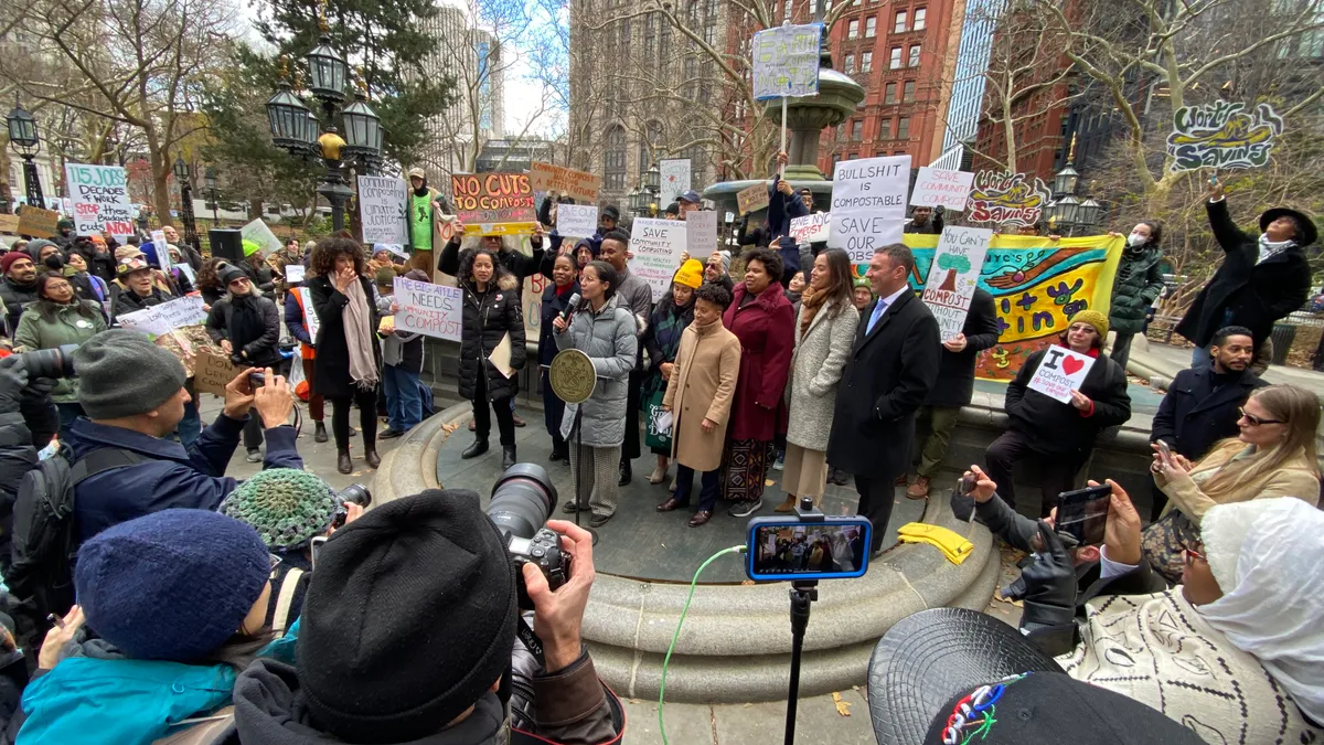 Nine people stand on a concrete dais in a park. Photographers snap their image in the foreground while ralliers hold signs with messages like "Bullshit is compostable" surround them.