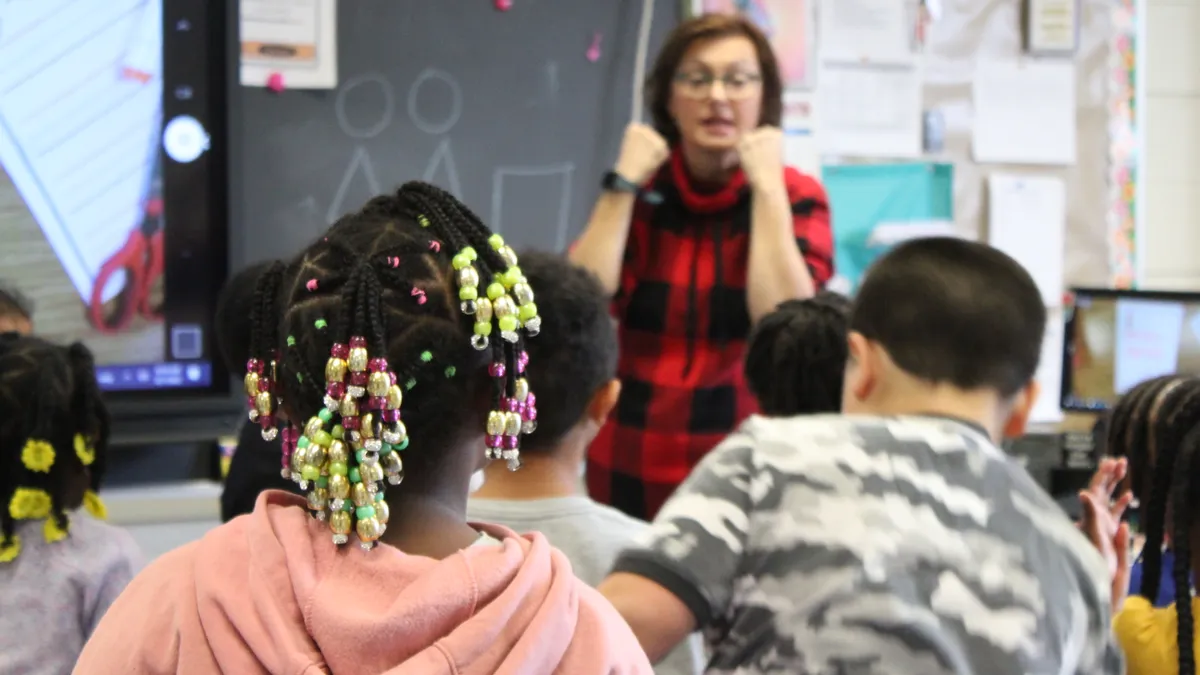 In the background is a teacher in a classroom leading a lesson. The background is slightly blurred and in the foreground are the backs of two students' heads.