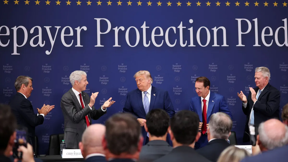 US President Donald Trump stands on stage while people around him applaud. The word "protection" can be seen behind him.