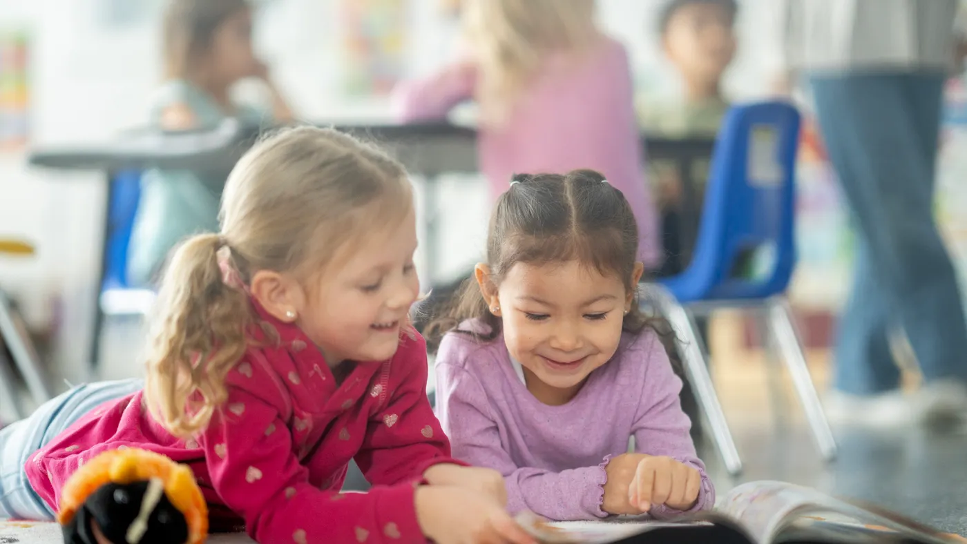 Two young students are on the floor in a classroom reading a book.