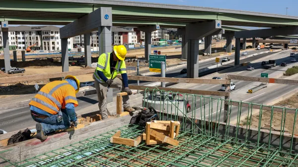 Two construction workers in hardhats with a highway behind them.