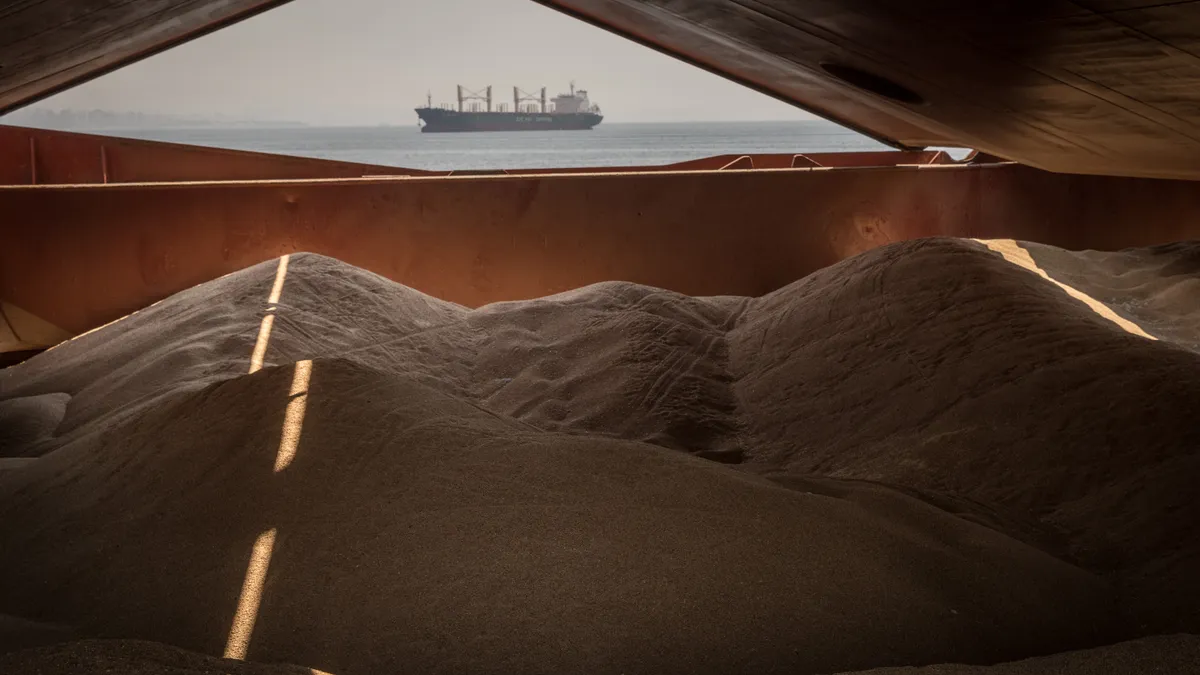 Piles of grain are seen from the perspective from inside a ship. A window shows a container ship in the background.