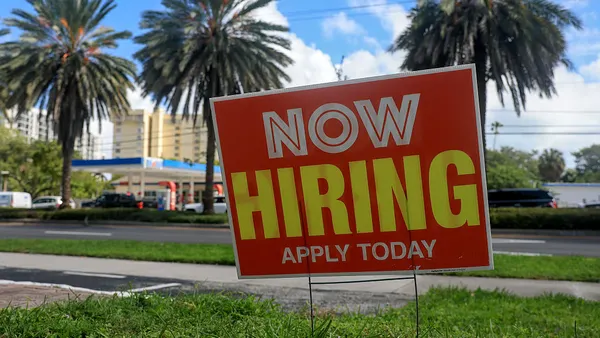 A lawn sign that reads "Now Hiring" is shown in the foreground, with lush palm trees in the background