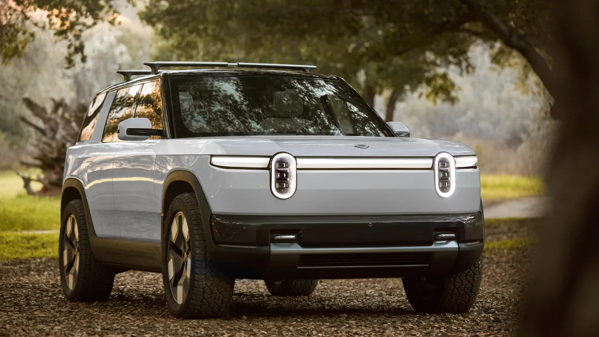 A white Rivvian R2 SUV parked outside under a tree with greenery in the background.