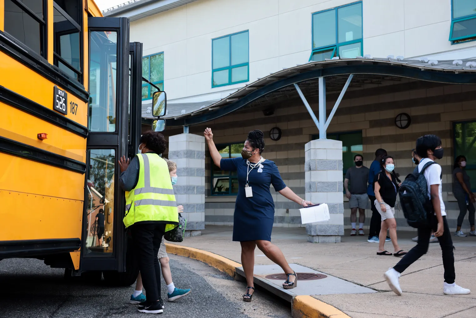 An adult stands outside near a yellow school bus parked alongside the curb near a school. Students are walking off the bus and toward the school.