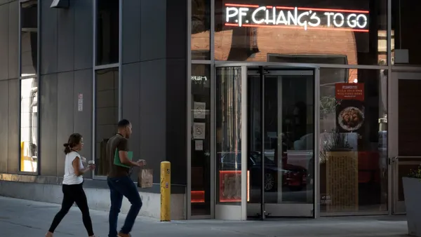 Two people walk by a P.F. Chang's To Go storefront. The building has a neon sign that reads "P.F. Chang's To Go".