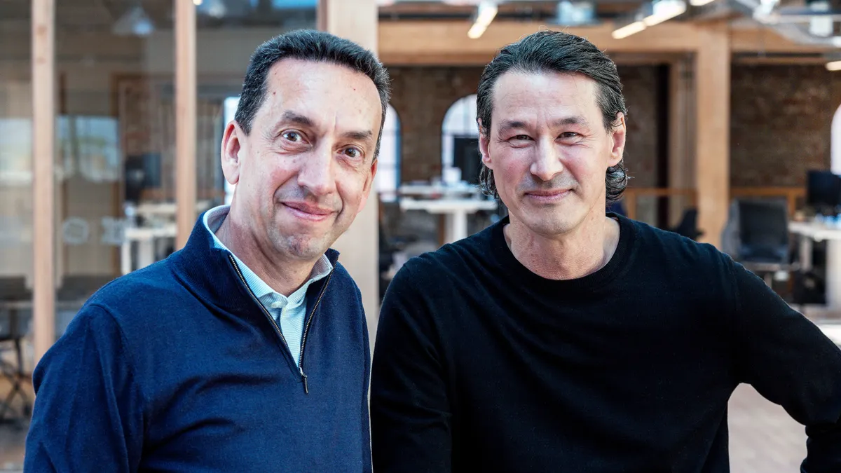 Jean-Christophe Flatin, in a navy blue sweater, stands next to Toni Peterson, in a black sweater, with an open office in the background.