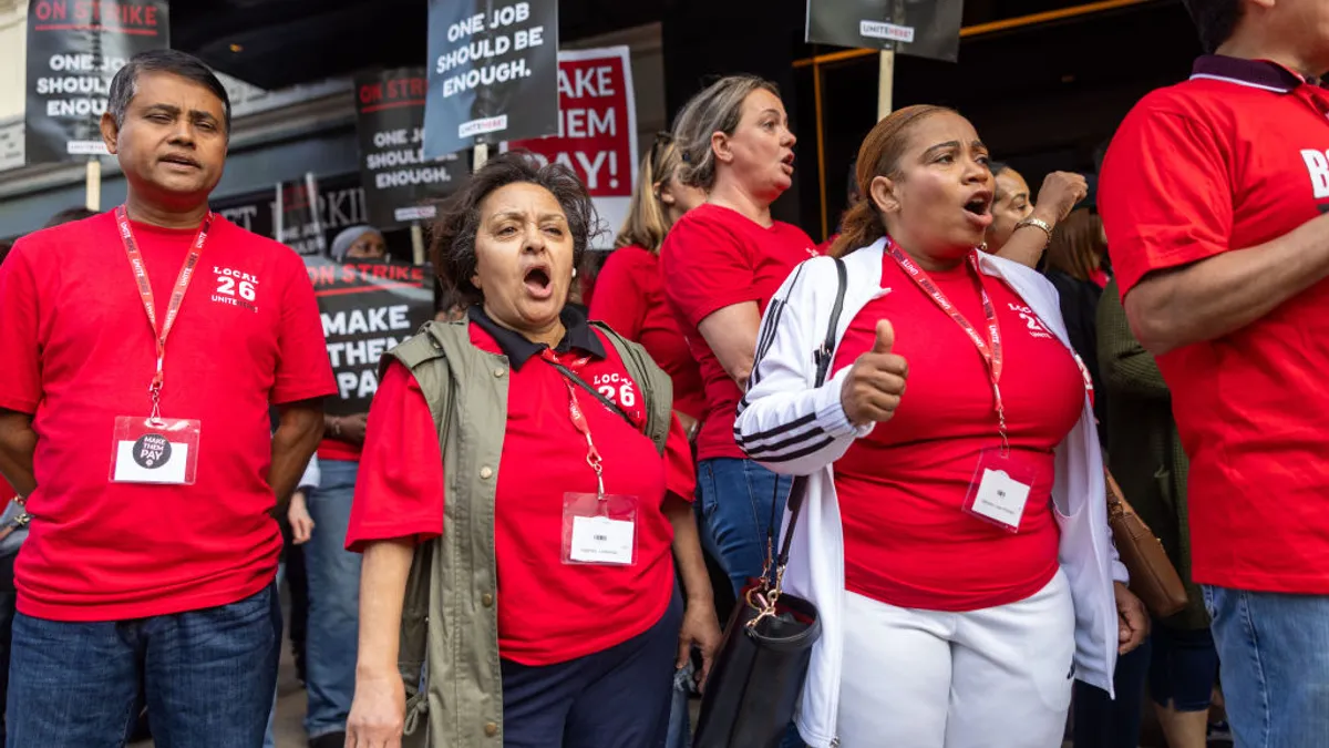 Workers stand together on a picket line and appear to be shouting.