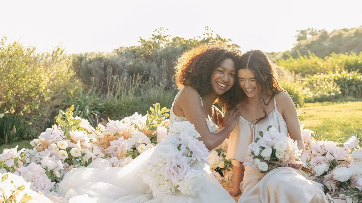 Two people in wedding dresses sit in a field of white silk flowers.