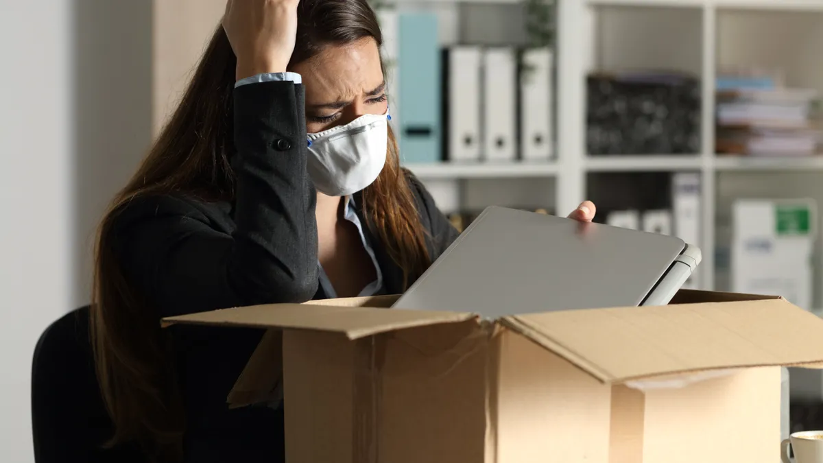 A woman in a mask looks upset and packs belongings into box.