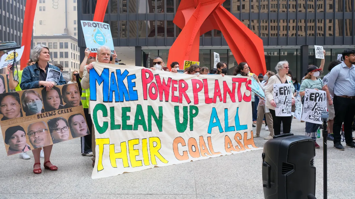 Protesters gather near a speaker holding signs. One sign has eight portraits and says "The Faces of Blews Creek," another says "MAKE POWER PLANTS CLEAN UP ALL THEIR COAL ASH."