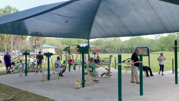 People using equipment and walking around in an outdoor fitness station covered with a large fabric shade with a grassy field behind them