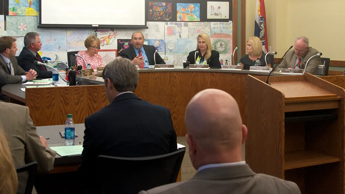 School board members sit during a meeting behind a podium with an audience looking toward them