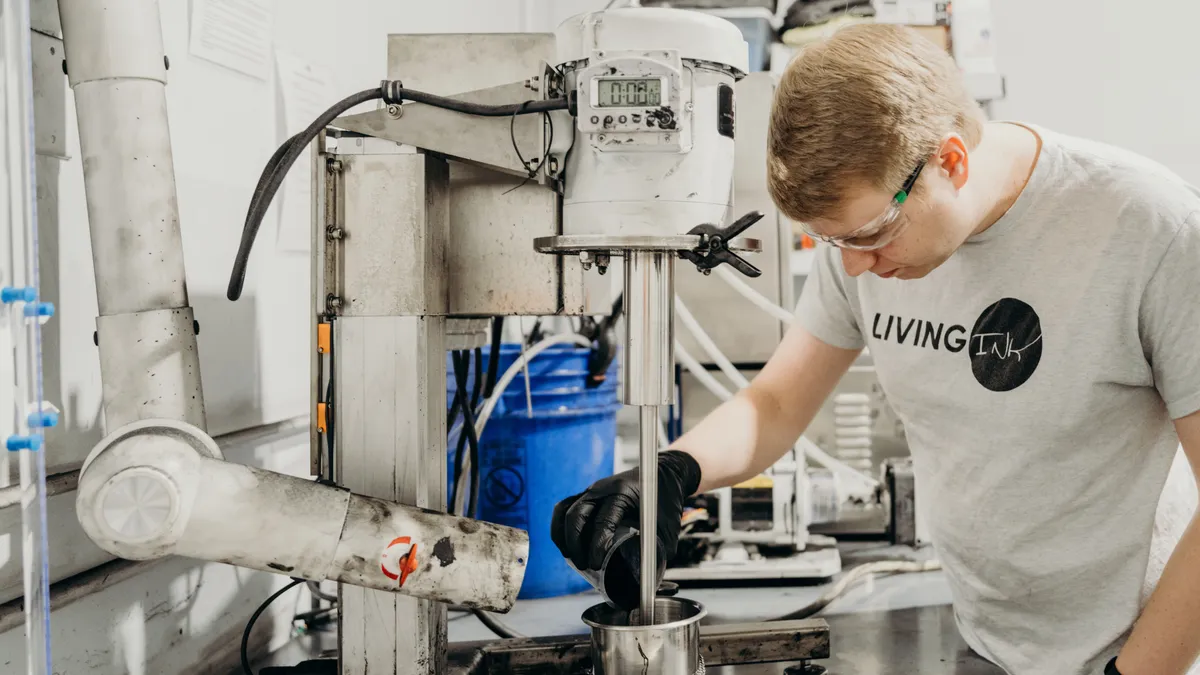 A person in a Living Ink t-shirt, wearing safety glasses and gloves, works with ink at an industrial station.