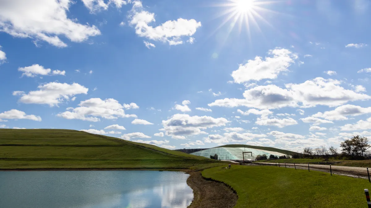 A retention pond in the foreground reflects a partly cloudy sky. Behind it are a grassy hill and a further hill covered in a tarp-like material.