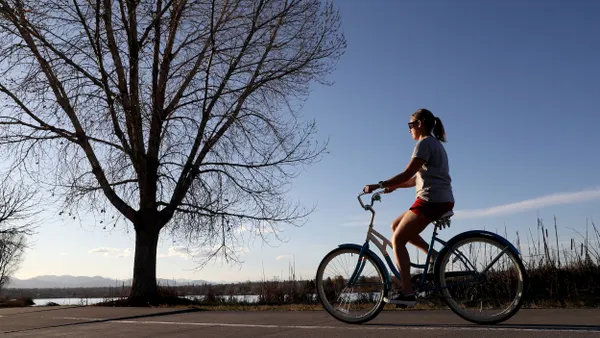 Woman in t-shirt and shots riding a bike along a paved path aside a large bare tree  with water and mountains in the background.