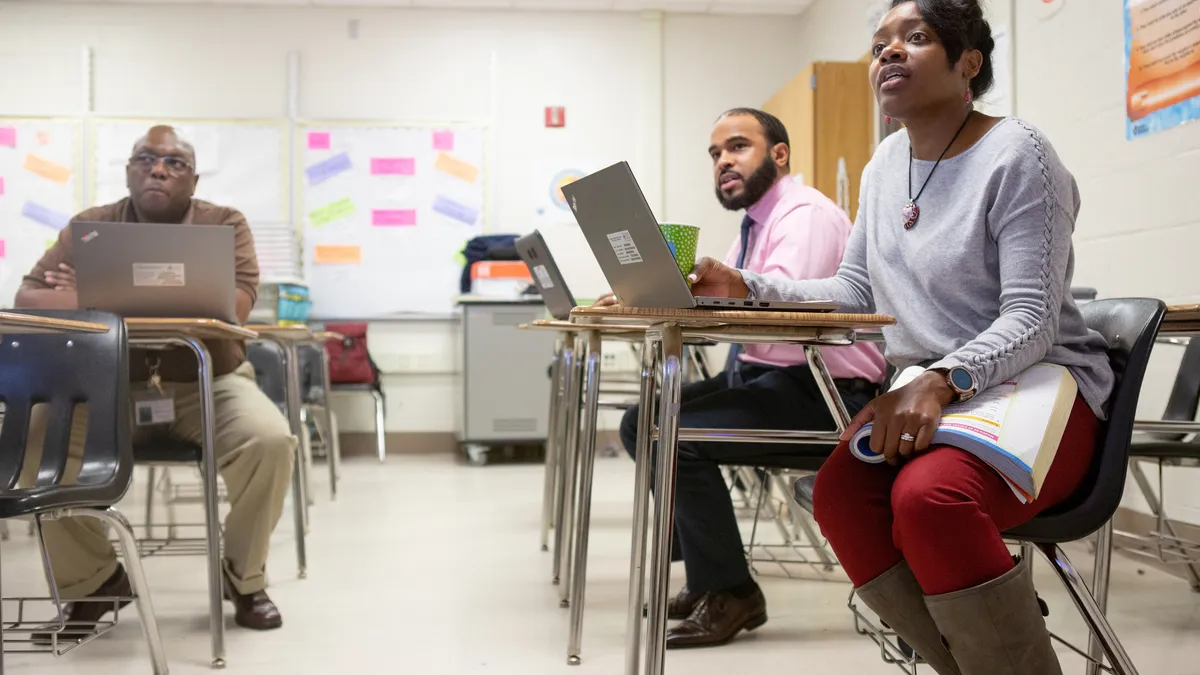 Three Black teachers sit at student desks in a school classroom during a professional development session.