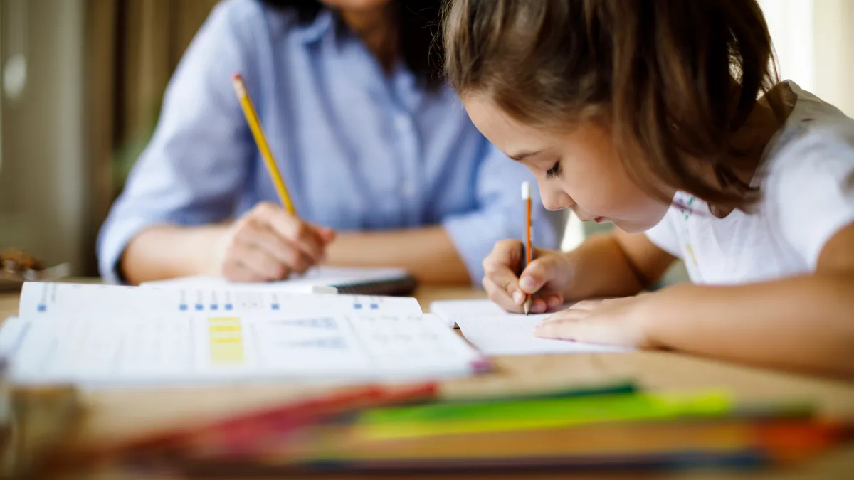 Student in foreground sits at a table with an adult. Both are holding pencils and there are workbooks on the table.