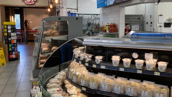 A photo of the open kitchen in a convenience store. The work area of the kitchen can be seen behind a glass cold case with sandwiches and other packaged foods.