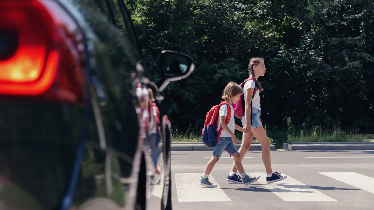 Two young children are crossing the street at a crosswalk while a car waits for them.