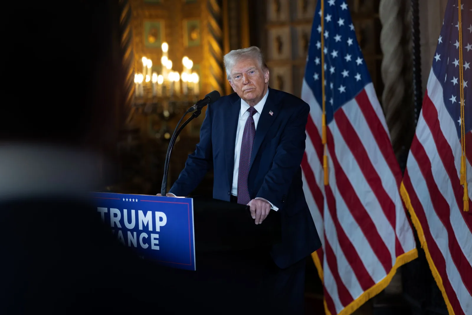 A picture of President-elect Donald Trump standing behind  a podium at a press conference with two American flags in the background.