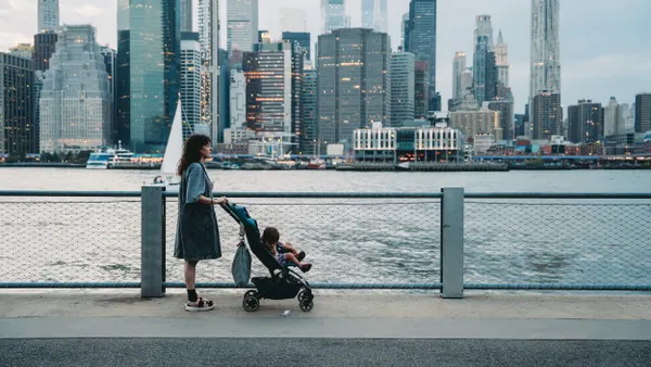 A person stands behind a baby in a stroller with water and city skyline in the background.